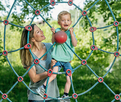 Kleiner blonder Junge mit Behinderung im Kletternetz auf Spielplatz, Mutter steht daneben.