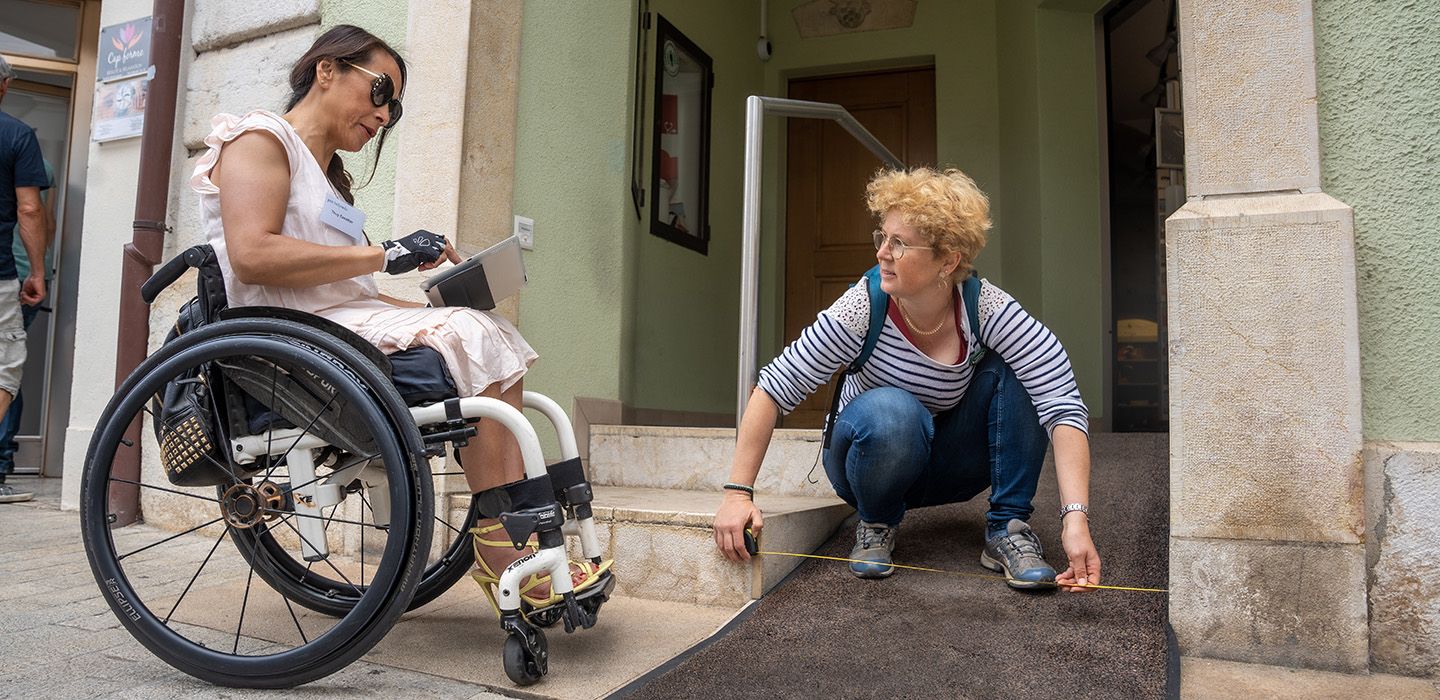 Zwei Personen (eine im Rollstuhl) vermessen einen Gebäude-Eingang