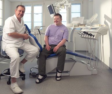 Ein Zahnarzt und ein Patient mit Down-Syndrom in einer Zahnarztpraxis lächeln in die Kamera.