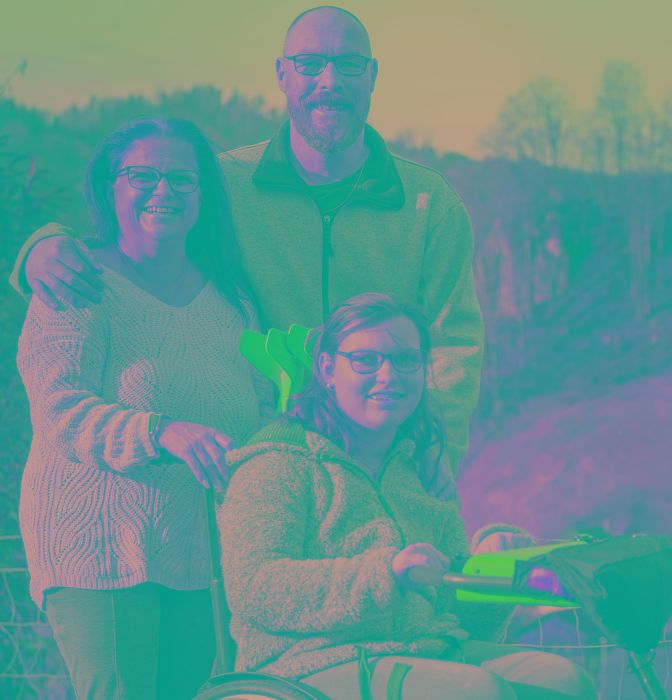 Jeune femme en fauteuil roulant devant un paysage de montagne, ses parents sont derrière elle.