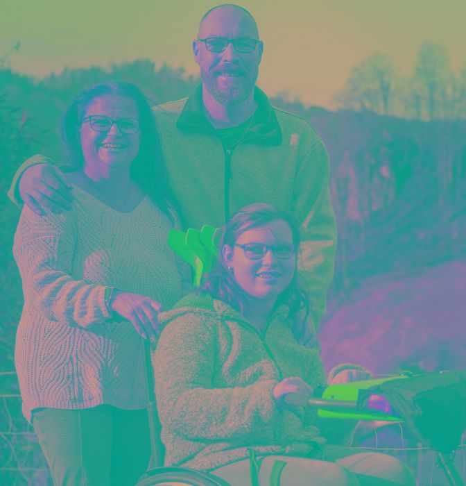 Junge Frau im Rollstuhl vor einer Berglandschaft, hinter ihr stehen ihre Eltern.