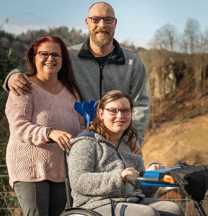 Jeune femme en fauteuil roulant devant un paysage de montagne, ses parents sont derrière elle.