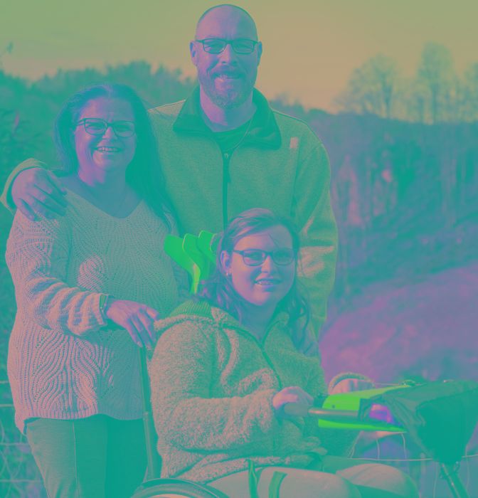 Giovane donna in sedia a rotelle davanti a un paesaggio montano, con i genitori in piedi dietro di lei.