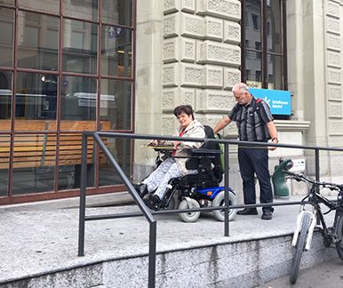 Frau im Rollstuhl mit Begleiter auf der Rampe zu einem öffentlichen Gebäude