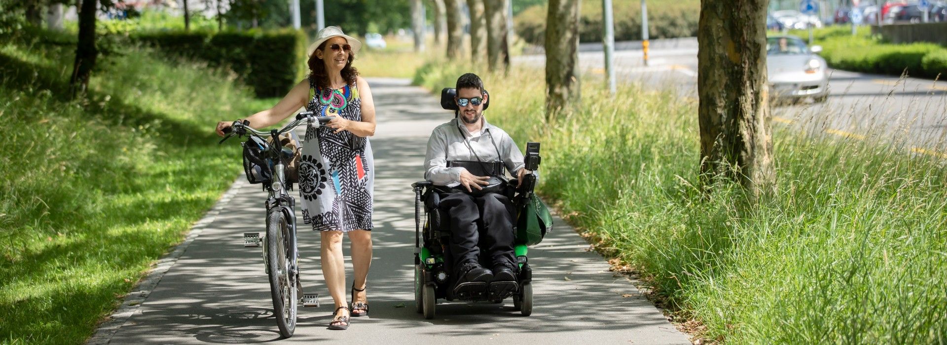 Ein junger Mann im Rollstuhl wird von einer Frau mit Velo begleitet
