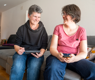 Zwei Freundinnen sitzen auf der Couch und lachen sich an.