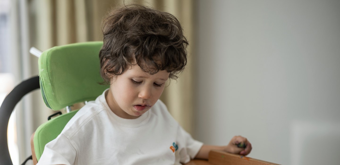 	Józef sitzt in einem grünen Therapiesitz am Tisch und hält einen Stift in der Hand.