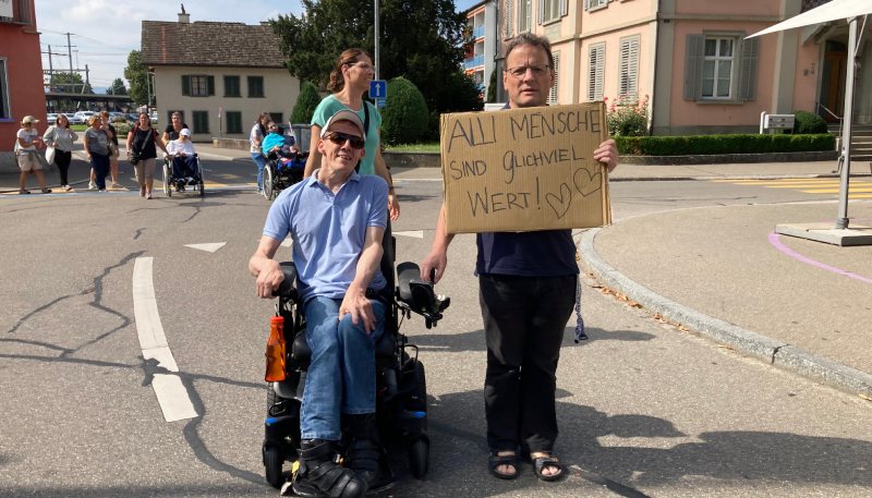 Ein Mann mit einem Schild neben einem Mann mit Rollstuhl beim Sternenmarsch in Weinfelden, im Hintergrund mehrere Personen