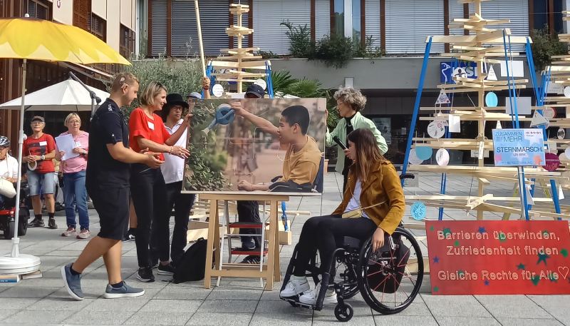 Mehrere Personen vor einem Bild bei der Kundgebung in Weinfelden, eine Person mit Rollstuhl