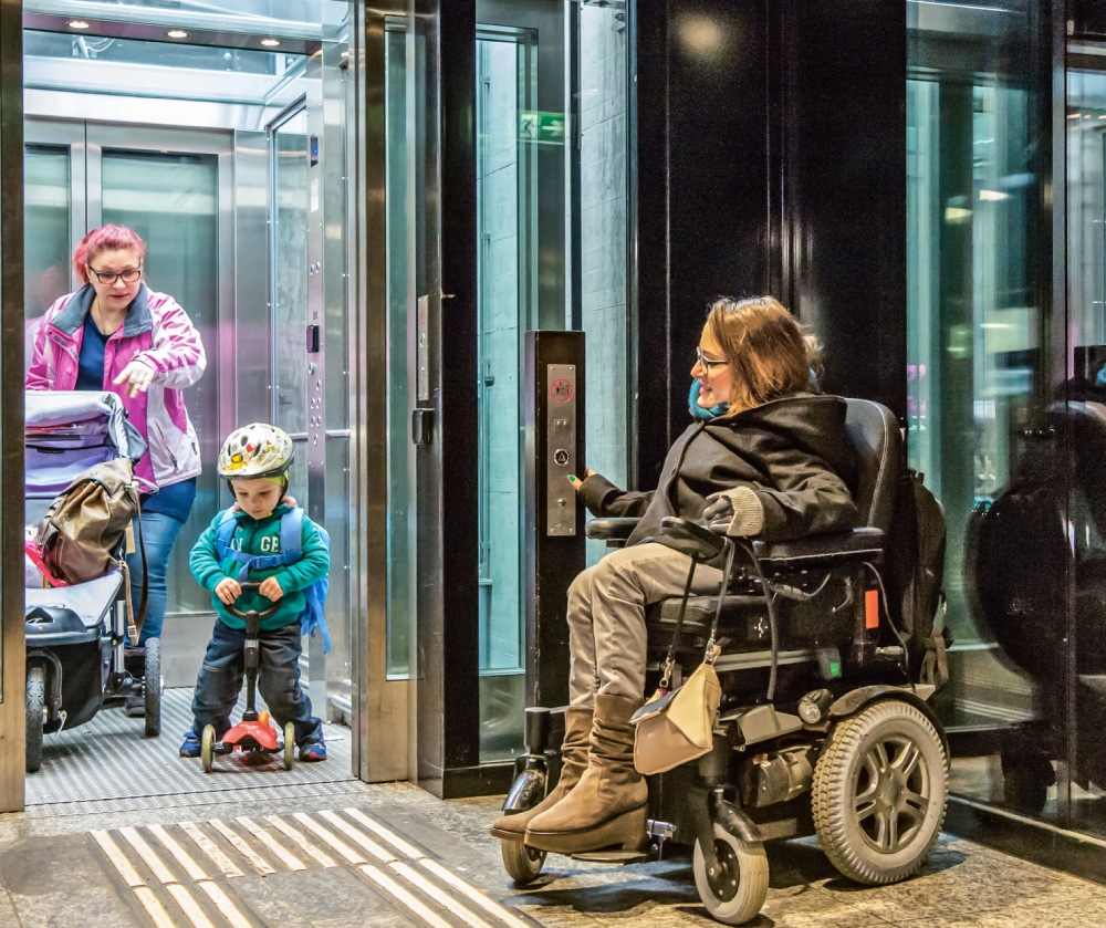Une femme avec une poussette et un jeune enfant dans un ascenseur, devant, une femme en fauteuil roulant.