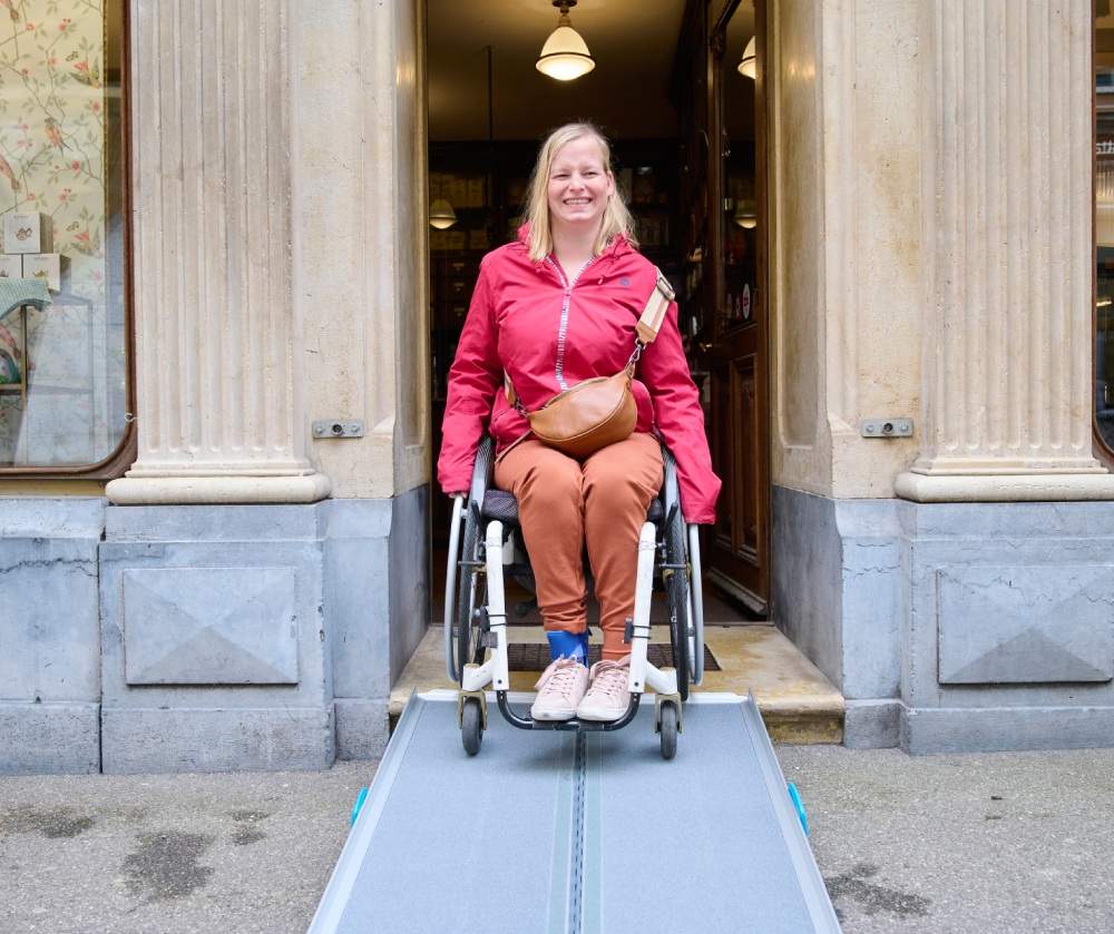 Frau im Rollstuhl fährt über eine Rampe aus einem Gebäude heraus.