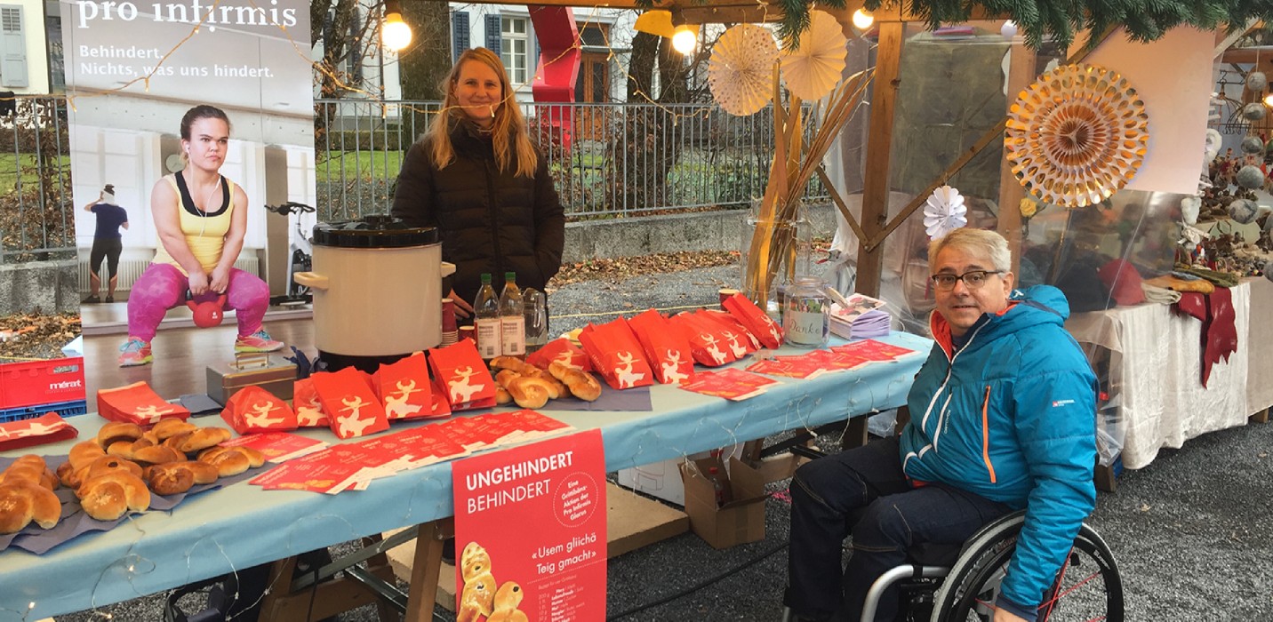 Bancarella con Grittibänz, una donna in piedi e un uomo in sedia a rotelle.