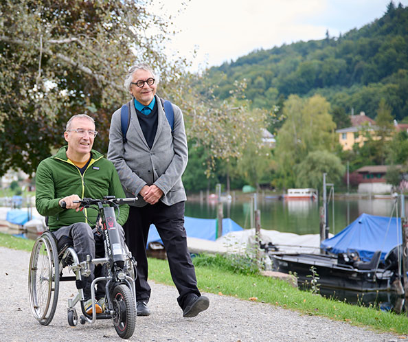 Un homme en chaise roulante se promène avec un autre le long d’un fleuve.