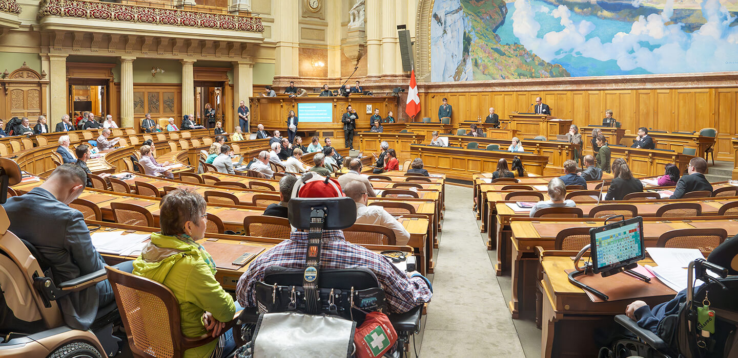 Teilnehmende der Behindertensession 2023 auf den Parlamentsplätzen im Nationalratssaal, darunter Personen im Rollstuhl.